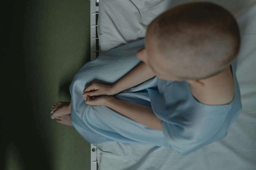 A cancer patient with a shaved head siting on a bed in the hospital
