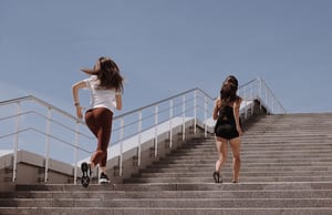 Two women running up outdoor stairs for fitness under a clear blue sky.